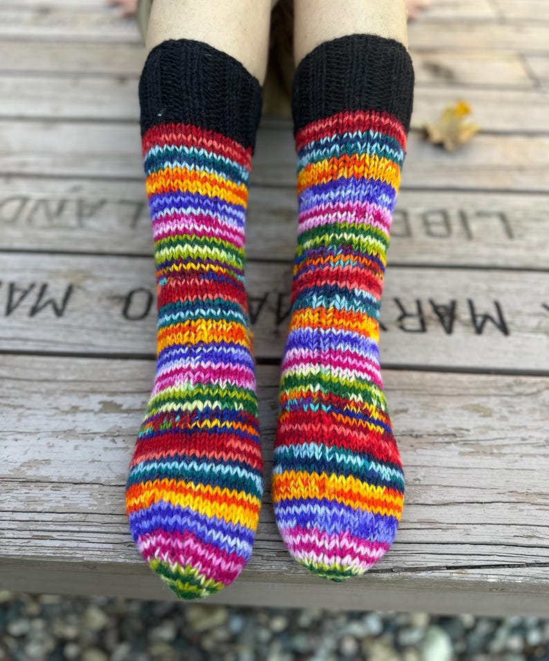 May include: A pair of hand-knitted socks featuring a vibrant, multi-colored striped pattern in shades of red, orange, yellow, green, blue, and purple. The socks have a solid black ribbed cuff at the top and a standard knit texture throughout.