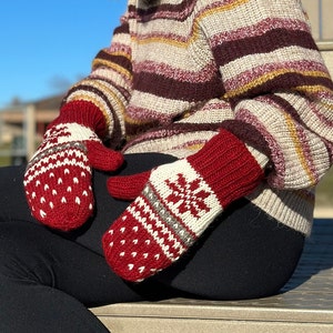 May include: A pair of red and white knit mittens with a snowflake pattern. The mittens are worn by a person wearing a striped sweater.