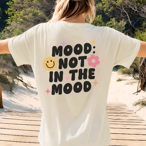 Pode incluir: Camiseta branca com um gr&aacute;fico preto que diz "Mood: Not in the Mood" com um rosto sorridente e uma flor rosa.