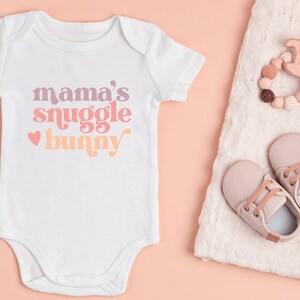 May include: White baby onesie with the text "mama's snuggle bunny" in pink, orange, and purple. A heart is to the left of the text. The onesie is on a pink background.