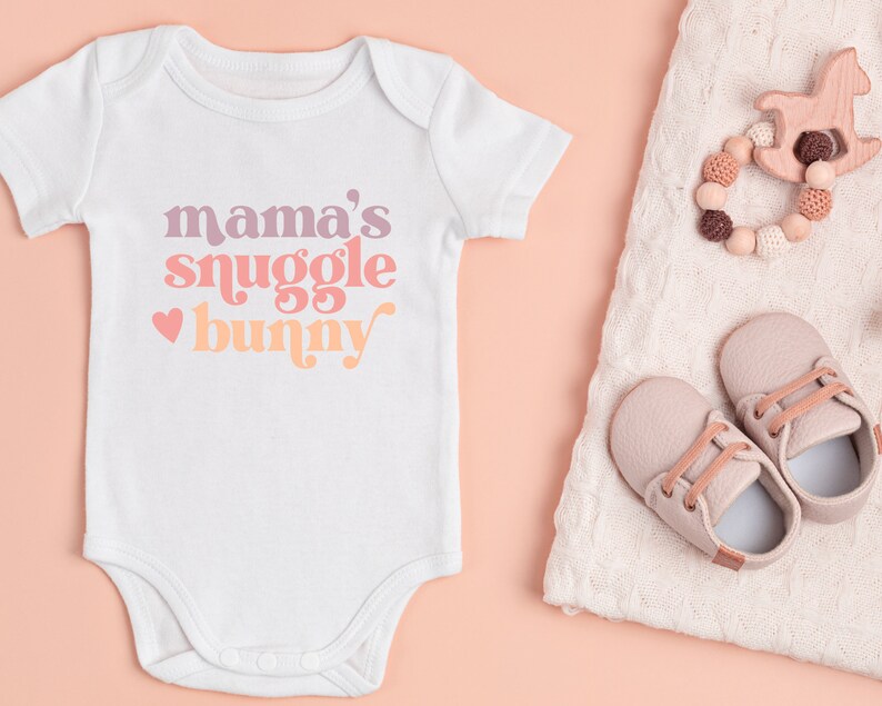 May include: White baby onesie with the text "mama's snuggle bunny" in pink, orange, and purple. A heart is to the left of the text. The onesie is on a pink background.