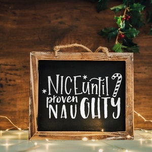 May include: A wooden framed chalkboard with the text "NICE until proven NAUGHTY" written in white chalk. The chalkboard is decorated with a candy cane and a star. The chalkboard is sitting on a table with string lights.
