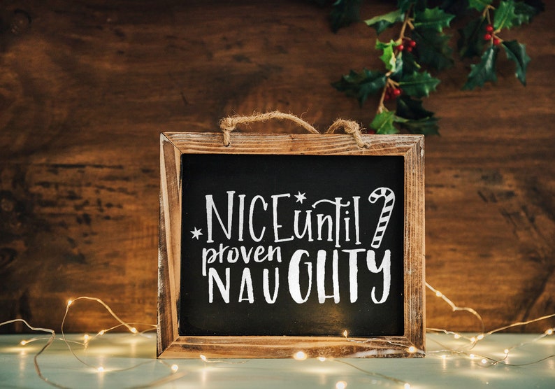 May include: A wooden framed chalkboard with the text "NICE until proven NAUGHTY" written in white chalk. The chalkboard is decorated with a candy cane and a star. The chalkboard is sitting on a table with string lights.