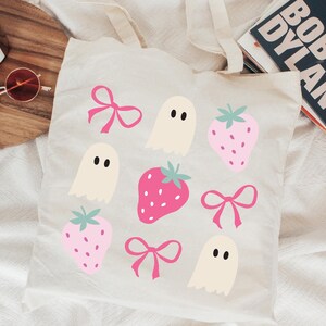 Puede incluir: Un bolso tote crema con un patr&oacute;n repetido de fresas rosas, lazos e ilustraciones de fantasmas. El bolso tiene dos asas y est&aacute; hecho de una tela suave. El dise&ntilde;o es caprichoso y juguet&oacute;n, ideal para llevar art&iacute;culos cotidianos.