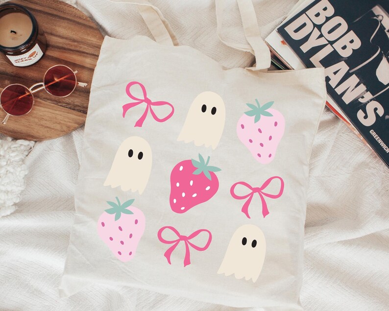 Puede incluir: Un bolso tote crema con un patr&oacute;n repetido de fresas rosas, lazos e ilustraciones de fantasmas. El bolso tiene dos asas y est&aacute; hecho de una tela suave. El dise&ntilde;o es caprichoso y juguet&oacute;n, ideal para llevar art&iacute;culos cotidianos.