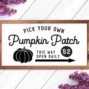 K&ouml;nnte beinhalten: Ein wei&szlig;es Schild mit einem braunen Holzrahmen, das einen K&uuml;rbis-Patch bewirbt. Das Schild lautet "Pick Your Own Pumpkin Patch" mit einer schwarzen Silhouette eines K&uuml;rbisses und einem Pfeil, der nach rechts zeigt. Der Text "This Way Open Daily" und "2&euro;" steht ebenfalls auf dem Schild.