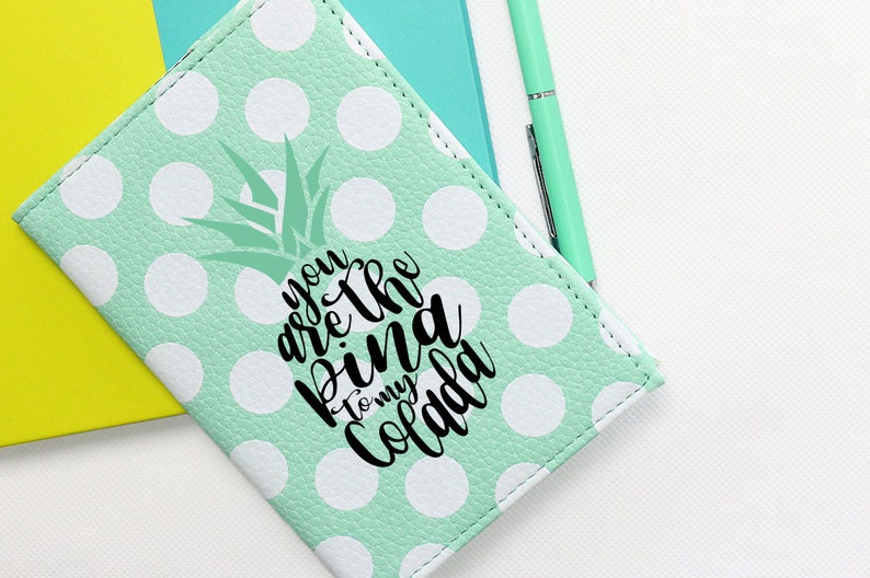 May include: A mint green passport holder with white polka dots and a pineapple graphic. The text "You are the pina to my colada" is printed on the holder.