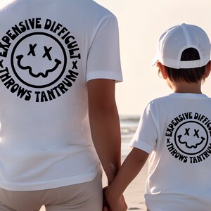 May include: White t-shirts with a black circle graphic on the back. The graphic features a smiley face with an 'X' for eyes and the text 'Expensive Difficult Throws Tantrum'.