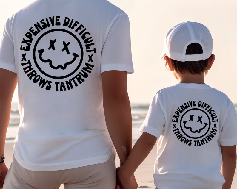 May include: White t-shirts with a black circle graphic on the back. The graphic features a smiley face with an 'X' for eyes and the text 'Expensive Difficult Throws Tantrum'.