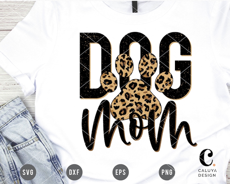 May include: A white t-shirt with black text that says "Dog Mom" with a leopard print paw print in the middle of the text.