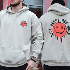 May include: A light beige hoodie with a black and red graphic on the front and back. The graphic is a dripping smiley face with the text "DADA. DADDY. DAD. BRUH." around it.