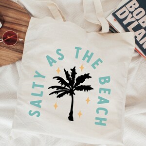 May include: Cream-colored tote bag with the text "SALTY AS THE BEACH" in turquoise and a black palm tree graphic. The design includes yellow star accents, perfect for beach lovers.