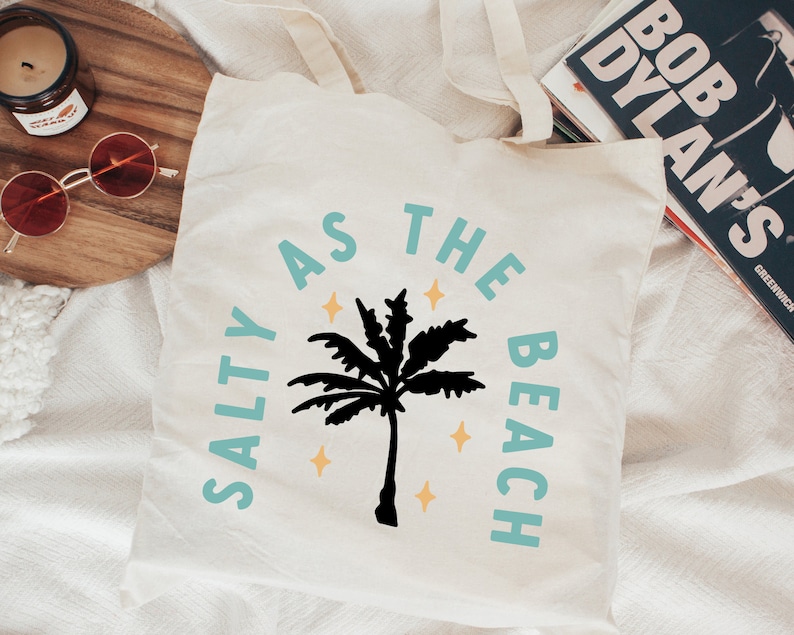 May include: Cream-colored tote bag with the text "SALTY AS THE BEACH" in turquoise and a black palm tree graphic. The design includes yellow star accents, perfect for beach lovers.
