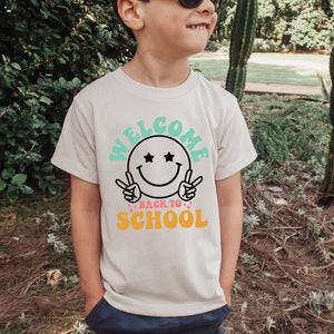 May include: A white t-shirt with a graphic design featuring a smiling face with stars for eyes and the text "Welcome Back to School" in colorful letters.