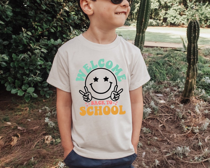 May include: A white t-shirt with a graphic design featuring a smiling face with stars for eyes and the text "Welcome Back to School" in colorful letters.