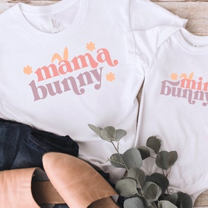 May include: Two white t-shirts with a pink, orange, and yellow graphic design. The larger shirt says "mama bunny" and the smaller shirt says "mini bunny".