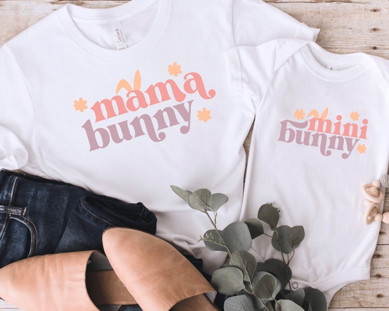 May include: Two white t-shirts with a pink, orange, and yellow graphic design. The larger shirt says "mama bunny" and the smaller shirt says "mini bunny".