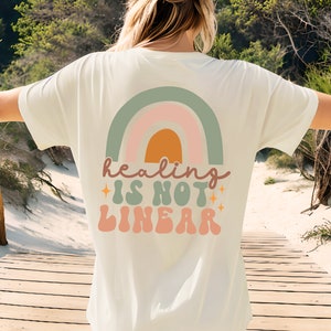 Op de afbeelding: Een wit T-shirt met een regenbooggrafiek en de tekst "healing is not linear" in een retro-lettertype.