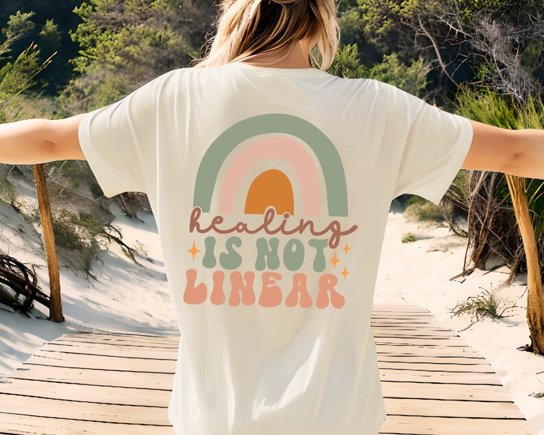 Op de afbeelding: Een wit T-shirt met een regenbooggrafiek en de tekst "healing is not linear" in een retro-lettertype.
