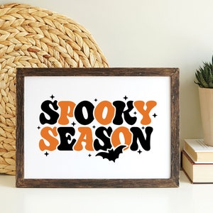 May include: A framed print with the text "Spooky Season" in orange and black retro font. A black bat silhouette is below the text.