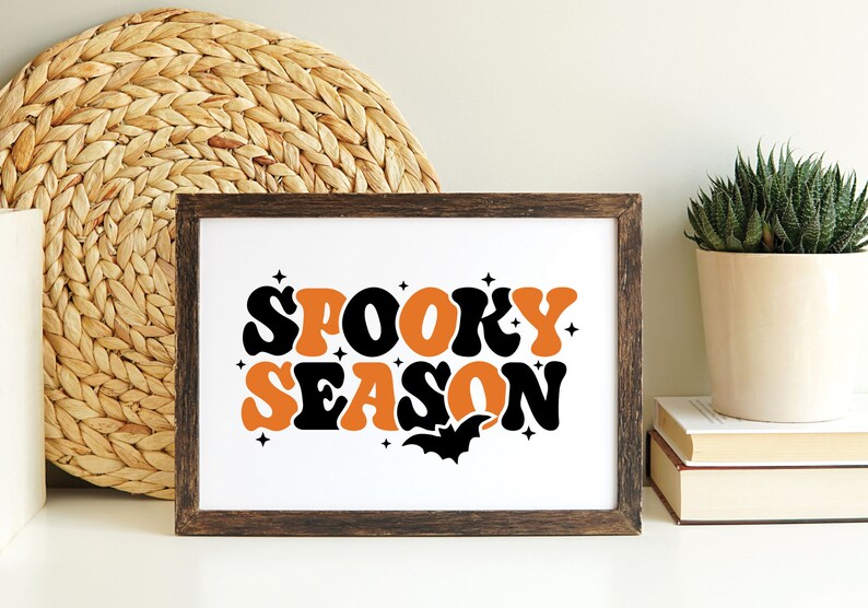 May include: A framed print with the text "Spooky Season" in orange and black retro font. A black bat silhouette is below the text.