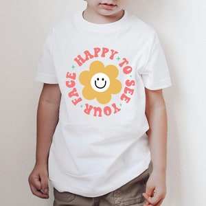 May include: White t-shirt with a pink and yellow flower graphic and the text "Happy to see your face".