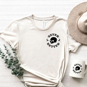Pu&ograve; includere: Una maglietta beige con una grafica di teschio nero e il testo "NEVER BETTER" in un cerchio. &Egrave; mostrata anche una tazza bianca con lo stesso design.