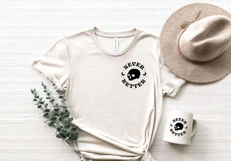 Pu&ograve; includere: Una maglietta beige con una grafica di teschio nero e il testo "NEVER BETTER" in un cerchio. &Egrave; mostrata anche una tazza bianca con lo stesso design.