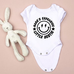 May include: White baby onesie with a black circle graphic that says "Mam's Expensive Little Bestie" with a smiley face and lightning bolts.