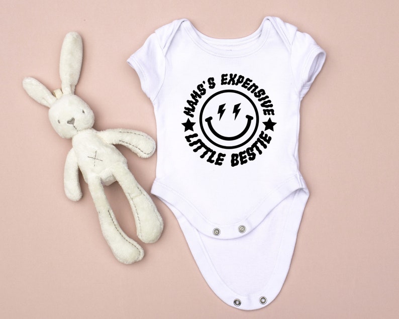 May include: White baby onesie with a black circle graphic that says "Mam's Expensive Little Bestie" with a smiley face and lightning bolts.