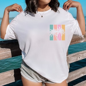 May include: White t-shirt with a colorful palm tree graphic. The graphic features a silhouette of palm trees in front of vertical stripes of teal, coral, pink, and yellow. The shirt is a casual, relaxed fit.