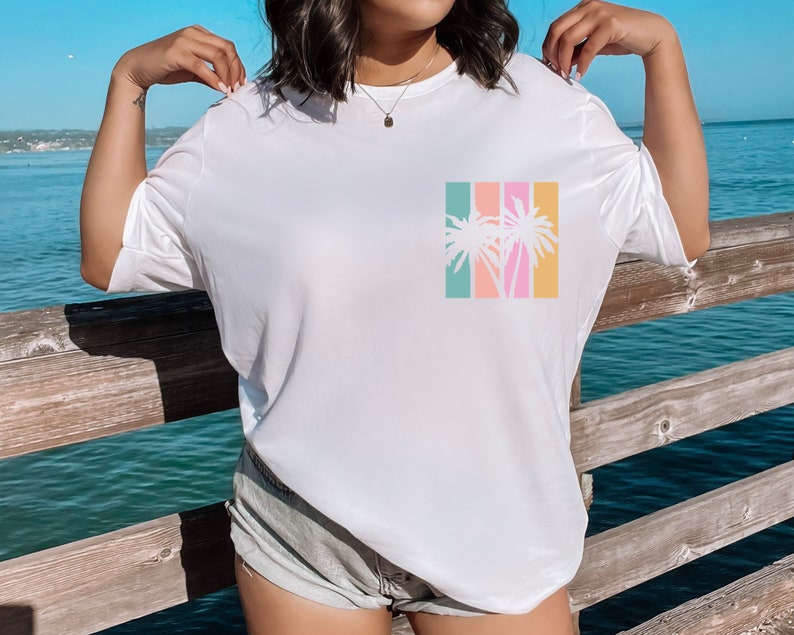 May include: White t-shirt with a colorful palm tree graphic. The graphic features a silhouette of palm trees in front of vertical stripes of teal, coral, pink, and yellow. The shirt is a casual, relaxed fit.