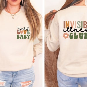 Op de afbeelding: Een cr&egrave;mekleurig sweatshirt met de tekst "Self Love Baby" op de voorkant en "Invisible illness Club" op de achterkant. De tekst is in een retro-stijl met kleurrijke letters en bloemen.