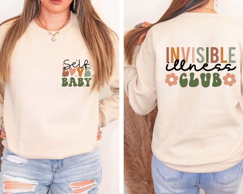 Op de afbeelding: Een cr&egrave;mekleurig sweatshirt met de tekst "Self Love Baby" op de voorkant en "Invisible illness Club" op de achterkant. De tekst is in een retro-stijl met kleurrijke letters en bloemen.