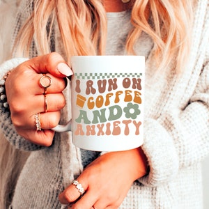 Op de afbeelding: Een witte keramische mok met een zwart-wit geruit ontwerp aan de bovenkant. De mok heeft een groen, bruin en oranje ontwerp met de tekst "I run on coffee and anxiety".