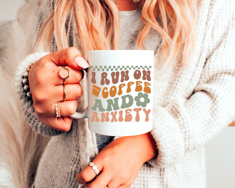 Op de afbeelding: Een witte keramische mok met een zwart-wit geruit ontwerp aan de bovenkant. De mok heeft een groen, bruin en oranje ontwerp met de tekst "I run on coffee and anxiety".