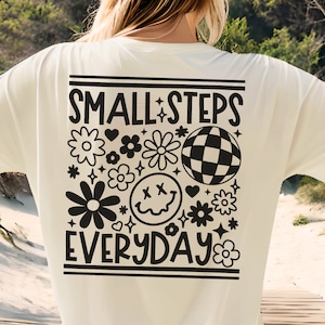 K&ouml;nnte beinhalten: Ein wei&szlig;es T-Shirt mit einem schwarzen Grafikdesign, auf dem "SMALL STEPS EVERYDAY" steht, umgeben von Blumen, einem Smiley und einer Discokugel.