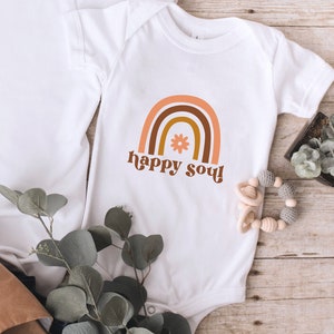 May include: White baby onesie with a brown, orange, and yellow rainbow graphic and the text "happy soul!" in a retro font. A small flower is in the center of the rainbow.