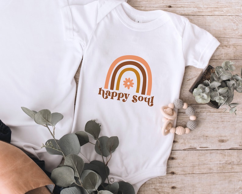 May include: White baby onesie with a brown, orange, and yellow rainbow graphic and the text "happy soul!" in a retro font. A small flower is in the center of the rainbow.