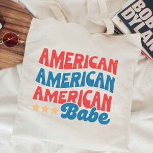 May include: White canvas tote bag with the text "American American American Babe" in red, blue, and white. Three gold stars are above the word "Babe".