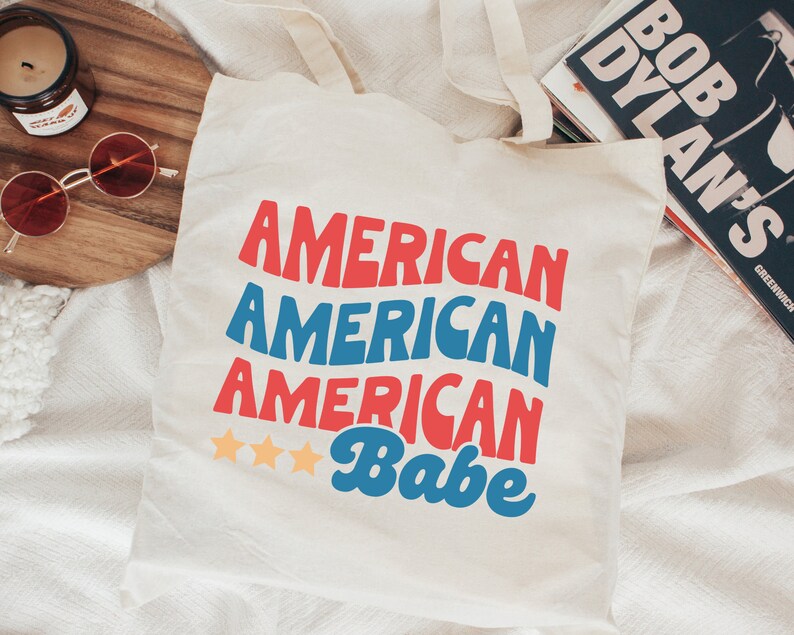 May include: White canvas tote bag with the text "American American American Babe" in red, blue, and white. Three gold stars are above the word "Babe".
