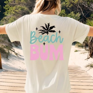 May include: Cream-colored t-shirt with the words "Beach Bum" in blue and pink, and a black palm tree graphic. The shirt is a relaxed fit, perfect for a day at the beach.