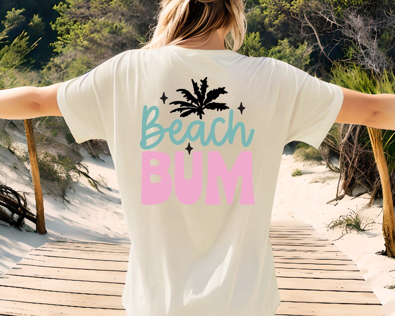 May include: Cream-colored t-shirt with the words "Beach Bum" in blue and pink, and a black palm tree graphic. The shirt is a relaxed fit, perfect for a day at the beach.