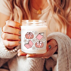 May include: A white candle with a pink and white design featuring pumpkins, books, and flowers. The candle is in a clear glass jar with a white label.