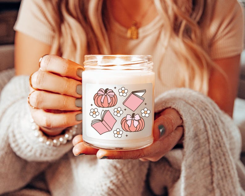 May include: A white candle with a pink and white design featuring pumpkins, books, and flowers. The candle is in a clear glass jar with a white label.