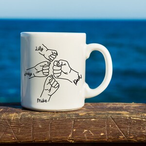 Pode incluir: Caneca de cer&acirc;mica branca com um desenho de linha preta de cinco m&atilde;os entrela&ccedil;adas. Os nomes Lily, Jay, Mike e Bud est&atilde;o escritos nas m&atilde;os.