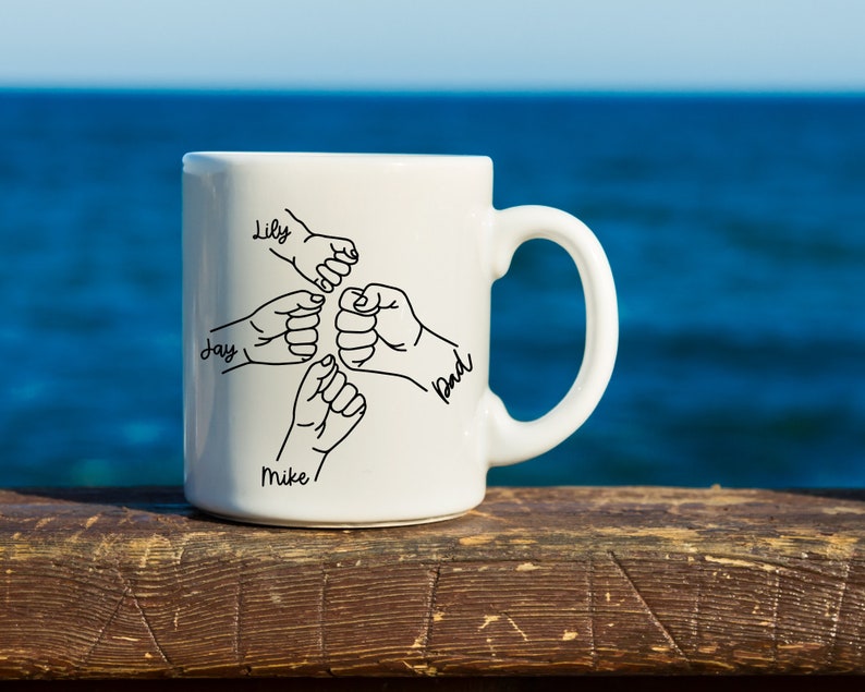 Pode incluir: Caneca de cer&acirc;mica branca com um desenho de linha preta de cinco m&atilde;os entrela&ccedil;adas. Os nomes Lily, Jay, Mike e Bud est&atilde;o escritos nas m&atilde;os.