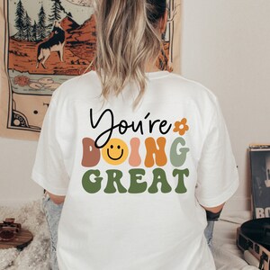 Op de afbeelding: Wit T-shirt met de tekst "You're doing great" in kleurrijke, retro-stijl letters. Een smiley is opgenomen in het woord "doing".