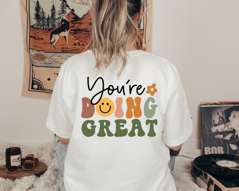 Op de afbeelding: Wit T-shirt met de tekst "You're doing great" in kleurrijke, retro-stijl letters. Een smiley is opgenomen in het woord "doing".