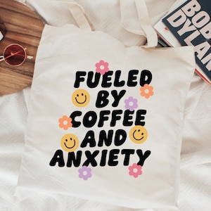 Pode incluir: Sacola de lona branca com o texto "Fueled by coffee and anxiety" em preto, com contorno branco, em uma fonte retr&ocirc;. O texto &eacute; cercado por pequenos rostos sorridentes amarelos e flores rosa e roxas.